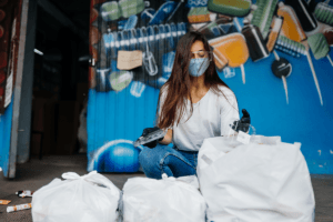 Woman wearing a mask sorting waste into white garbage bags in front of a blue mural.