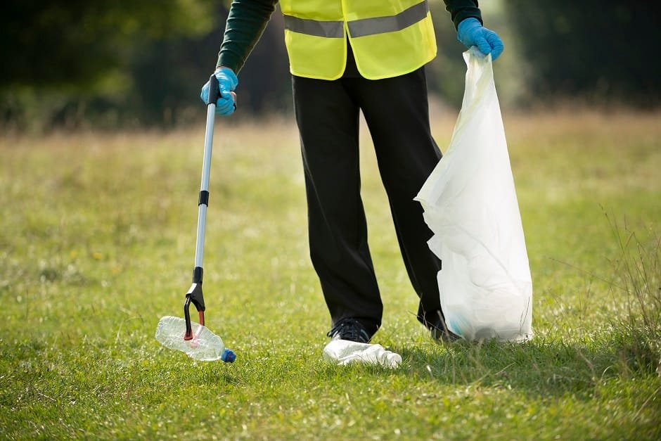 Person in safety vest picking up litter from grass using a grabber tool and trash bag.