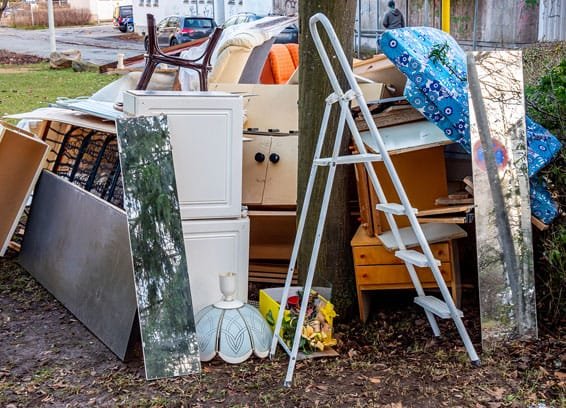 Discarded household furniture and items, including a ladder and cabinets, piled outdoors for removal.