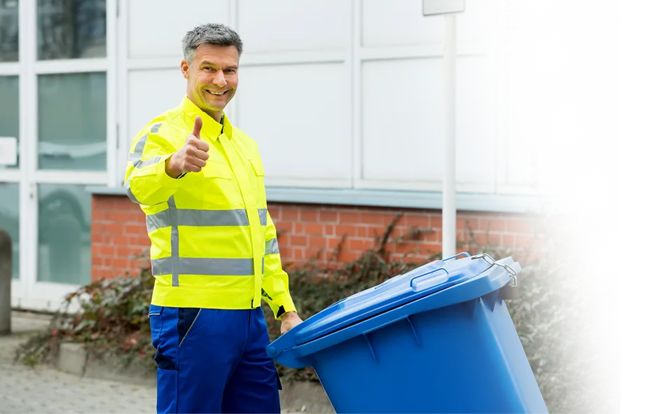 Smiling sanitation worker giving a thumbs-up beside a blue recycling bin outdoors.
